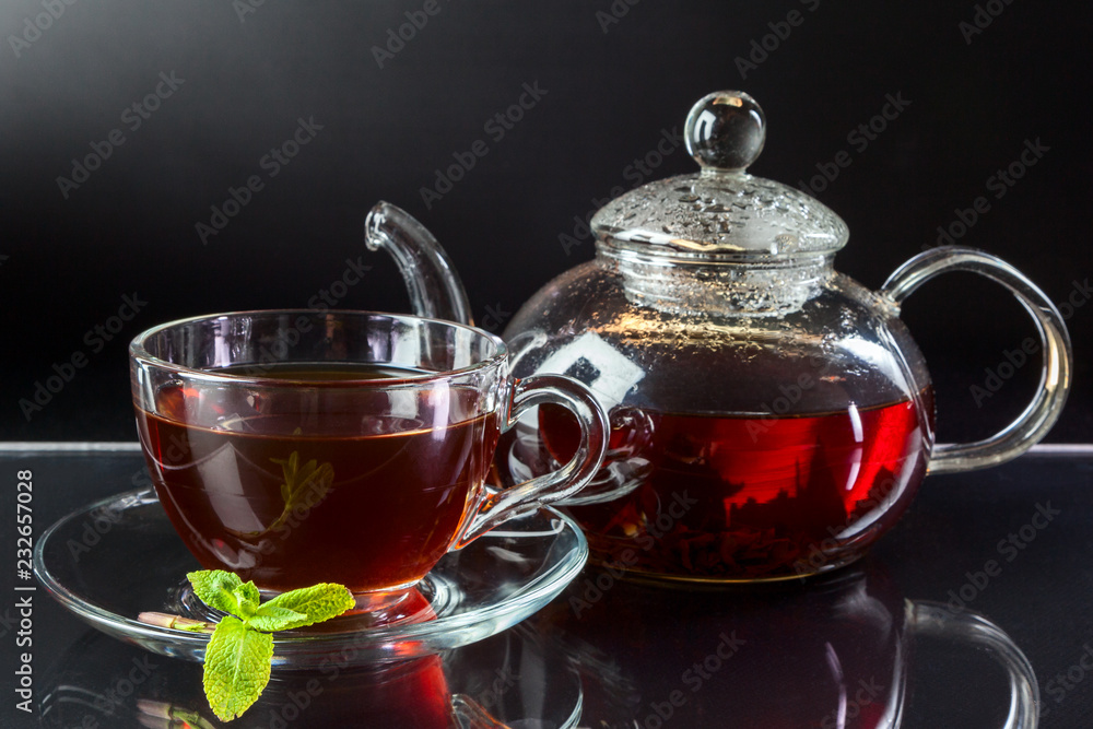 A transparent cup of black tea, a teapot and mint on a mirror table at a dark background A transparent cup of black tea, a teapot and mint on a mirror table at a dark background