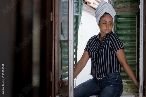 Chica joven con toalla de baño en la cabeza y gafas amarillas posando en la ventana de madera de una casa antigua