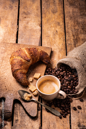 on the rustic wooden table, in the foreground, a cup of espresso coffee, a croissant with cereals, and roasted coffee beans.