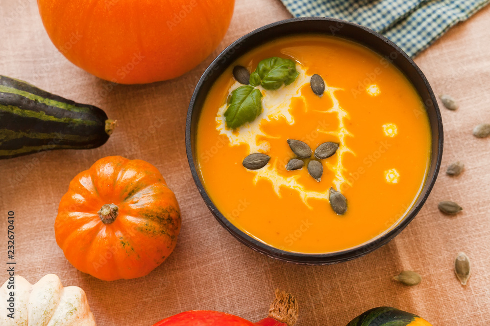 Pumpkin soup puree with cream and pumpkin seeds on the background of whole pumpkins of different colors. The surface of the textile Pumpkin soup puree with cream and pumpkin seeds on the background of whole pumpkins of different colors. The surface of the textile
