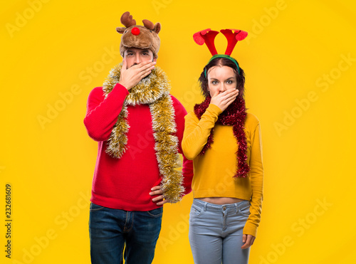 Couple dressed up for the christmas holidays covering mouth with hands for saying something inappropriate on yellow background