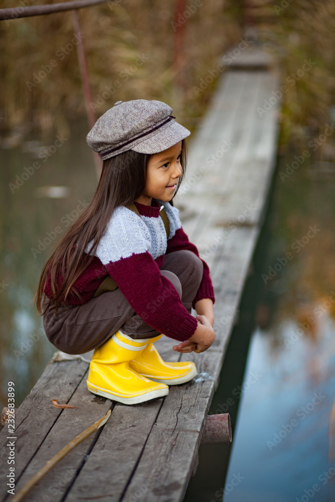 Obraz premium Child girl sits on wooden bridge and smiles on background of river.