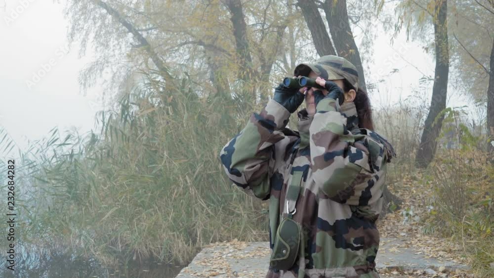 A beautiful Woman in army Camouflage Examines the Surroundings with Binoculars Standing on The River Bank on a Foggy Morning A beautiful Woman in army Camouflage Examines the Surroundings with Binoculars Standing on The River Bank on a Foggy Morning