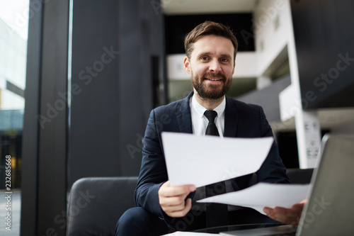 Happy bearded businessman with papers looking at you while working in modern office