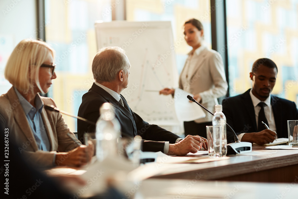 Mature delegate in formalwear paying attention to what young speaker by whiteboard saying Mature delegate in formalwear paying attention to what young speaker by whiteboard saying