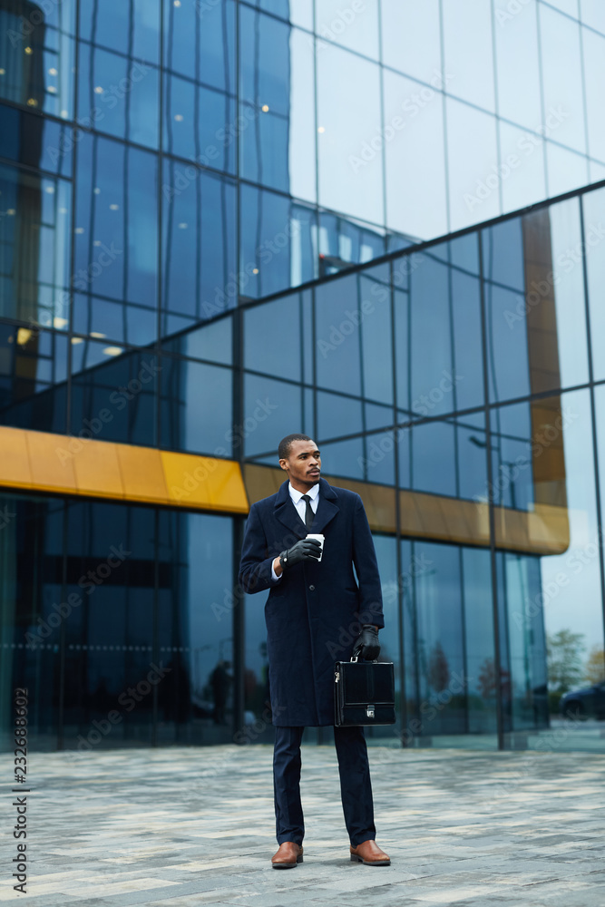 Young elegant African-american businessman with briefcase and drink standing by modern office center