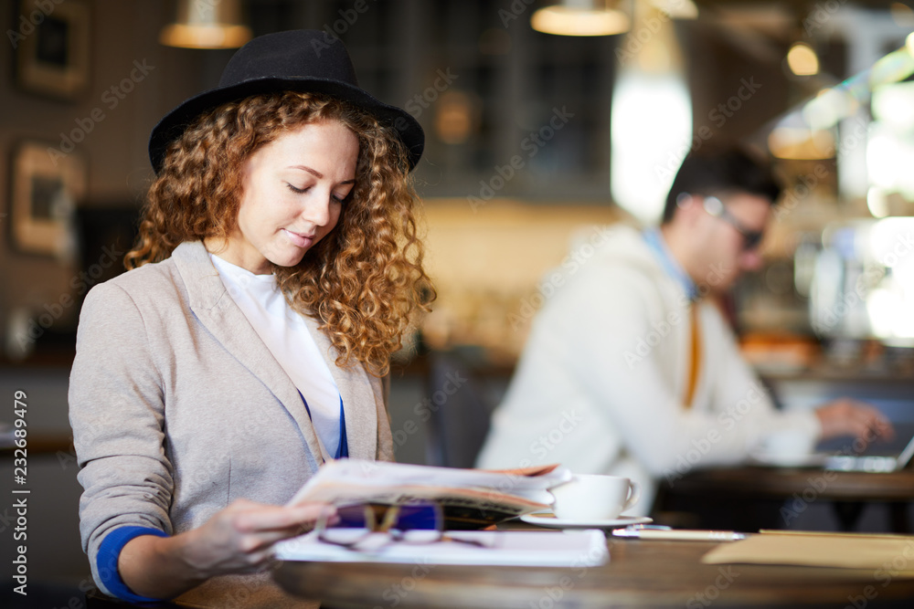 Fototapeta premium Young stylish wavy-haired businesswoman in hat reading the latest news in newspaper while sitting in cafe
