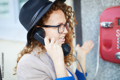Konstfotografi Young wavy-haired female in black hat and smart casual talking by payphone in bo