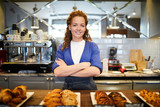Young cross-armed woman in workwear standing by counter with fresh pastry assortment