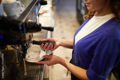 Young female barista making fresh aromatic coffee for one of clients while standing by coffee machine