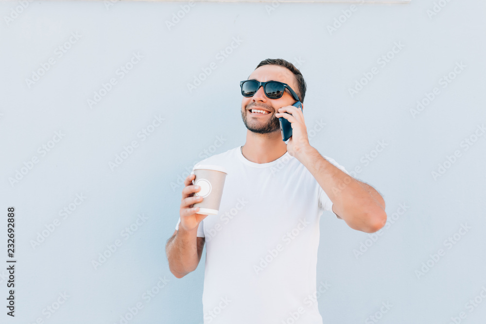 Happy male tourist satisfied with good connection in roaming talking with friend about vacations. Handsome man making mobile phone call while resting with coffee to go on blue background Happy male tourist satisfied with good connection in roaming talking with friend about vacations. Handsome man making mobile phone call while resting with coffee to go on blue background
