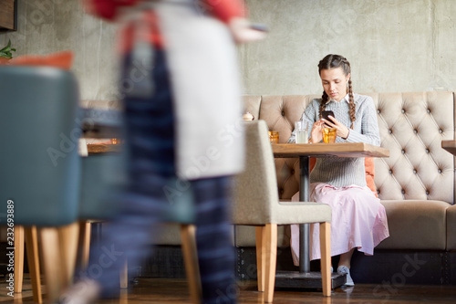 Casual girl with smartphone sitting on sofa by table and messaging while waiting for her order