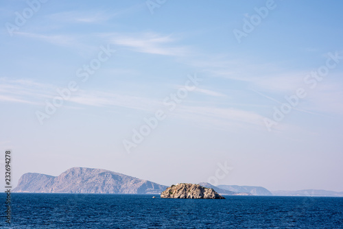 A small  rocky island juts out of a beautiful blue sea  in the Aegean Sea  near the Greek Island of Hydra.