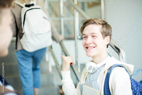 Happy guy pointing at girl with backpack moving upstairs while discussing her with friend