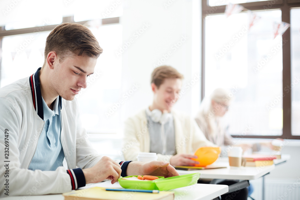 Obraz premium Casual guy and his groupmates having lunch at break from plastic containers while sitting in classroom