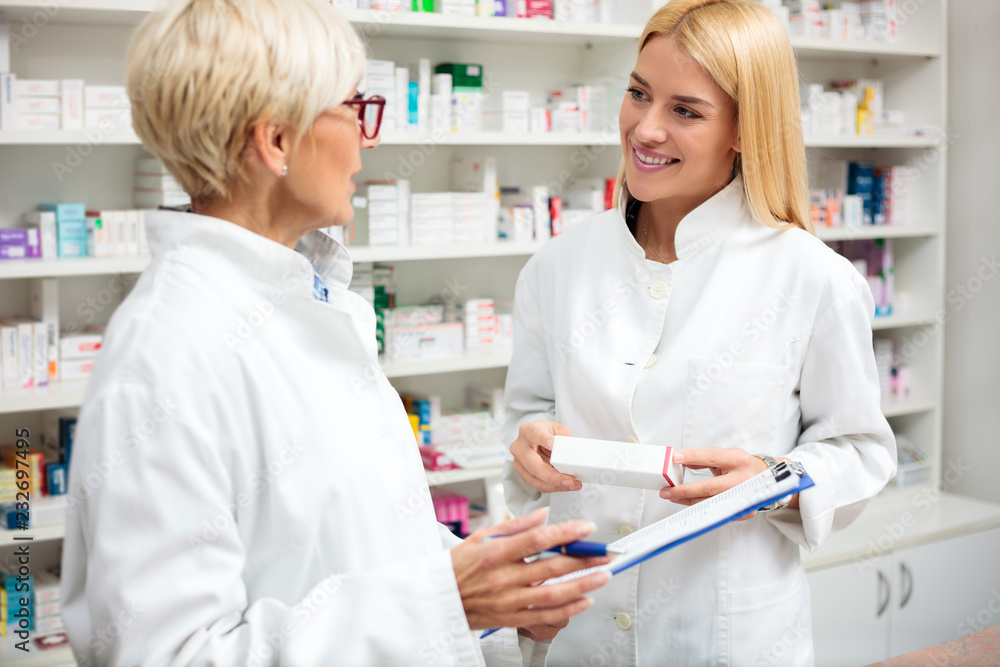 Happy young and mature blond haired female pharmacists standing in front of the shelves with medications, discussing. Writing on a clipboard and holding box of medications.