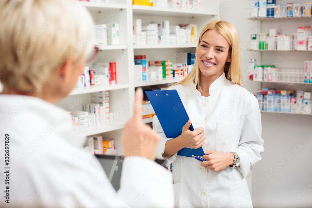 Mature and young female pharmacists working together, standing behind the counter. Young woman is holding a clipboard while her older colleague explains. Pharmaceutics and healthcare concept