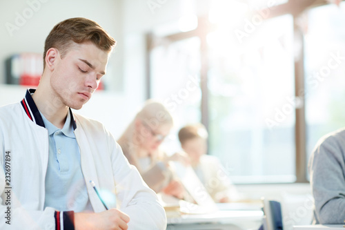 Serious guy in casualwear making notes or writing test at lesson among classmates