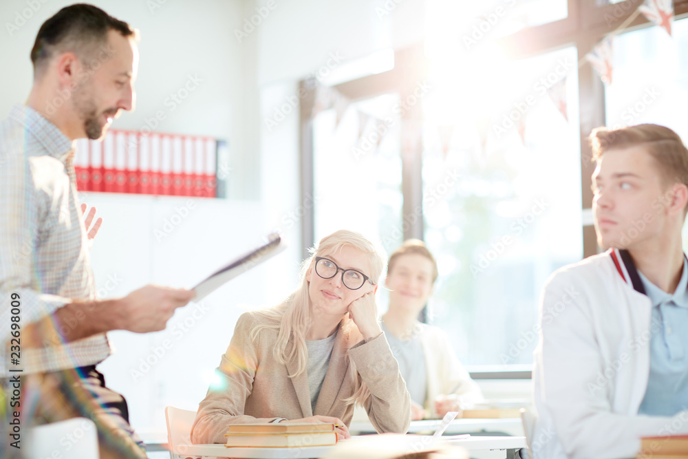 Obraz premium Happy blond girl in eyeglasses listening to what teacher saying while sitting at lesson