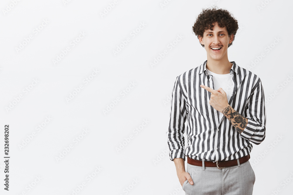 Studio shot of carefree happy and attractive hipser guy with dark curly har moustache and tattooed arm pointing at upper left corner holding hand in pants and smiling broadly with good intentions Studio shot of carefree happy and attractive hipser guy with dark curly har moustache and tattooed arm pointing at upper left corner holding hand in pants and smiling broadly with good intentions