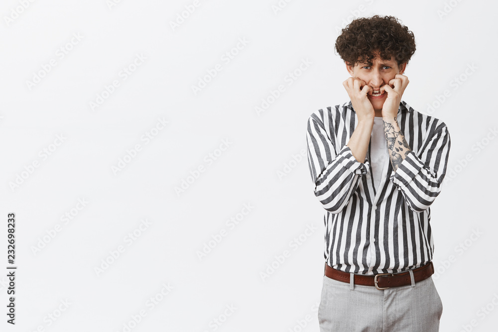 Portrait of frightened male coward with curly hair tattoos and moustache biting fingernails looking from under forehead scared and nervous panicking being terrified standing in striped shirt and pants Portrait of frightened male coward with curly hair tattoos and moustache biting fingernails looking from under forehead scared and nervous panicking being terrified standing in striped shirt and pants