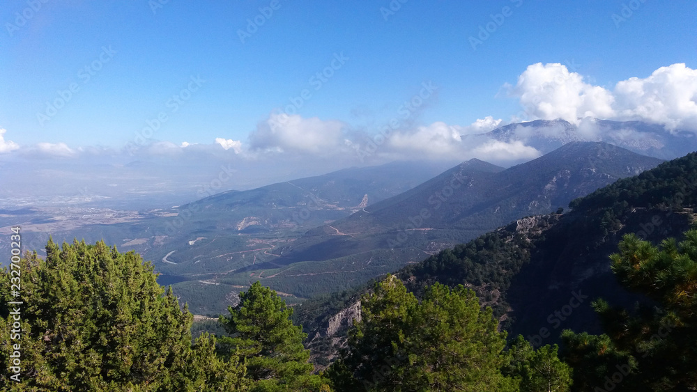Natural scenery at Denizli Kent Forest on a bright sunny morning, with green trees, mountaintops, bright blue skies and fluffy white clouds overlooking a green valley.