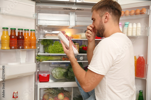 Young man with bad smelling meat near refrigerator in kitchen