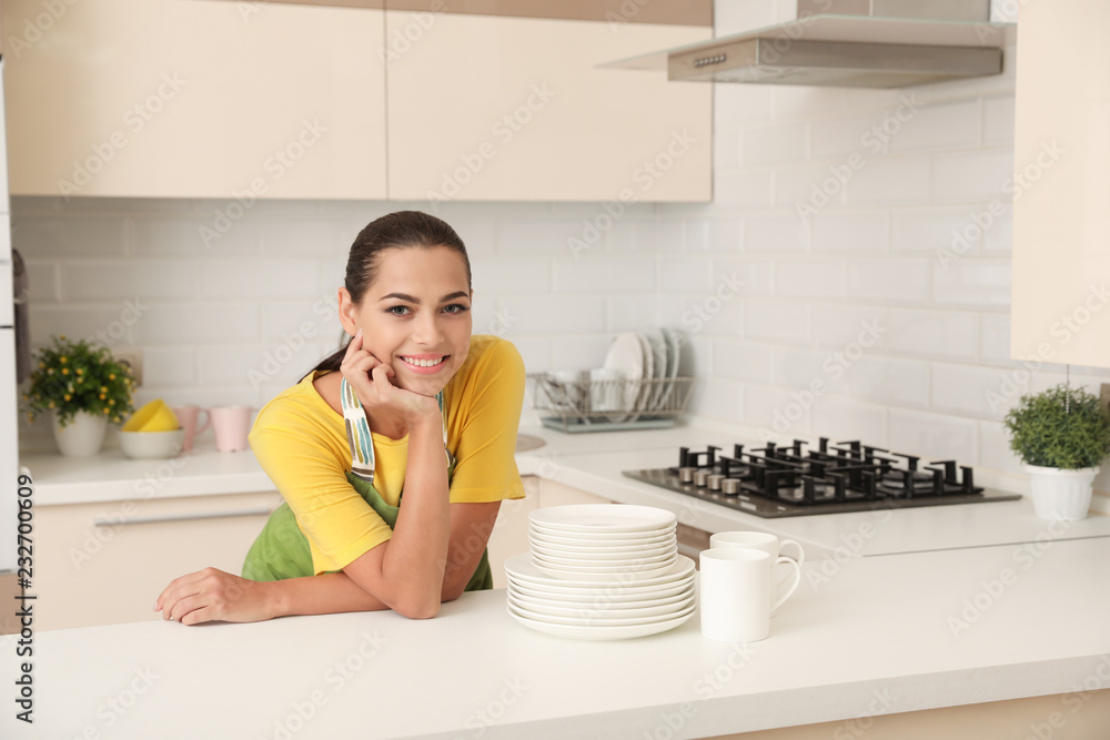 Beautiful young woman with clean dishes and cups at table in kitchen Beautiful young woman with clean dishes and cups at table in kitchen