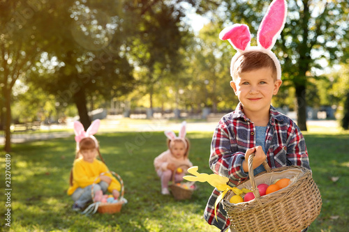 Cute little boy with bunny ears and basket of Easter eggs in park. Space for text