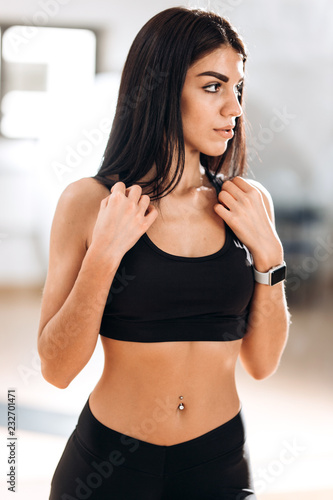 Fototapeta Naklejka Na Ścianę i Meble -  Beautiful slim girl dressed in black sportswear is standing in the gym in the fitness room next to the mirror