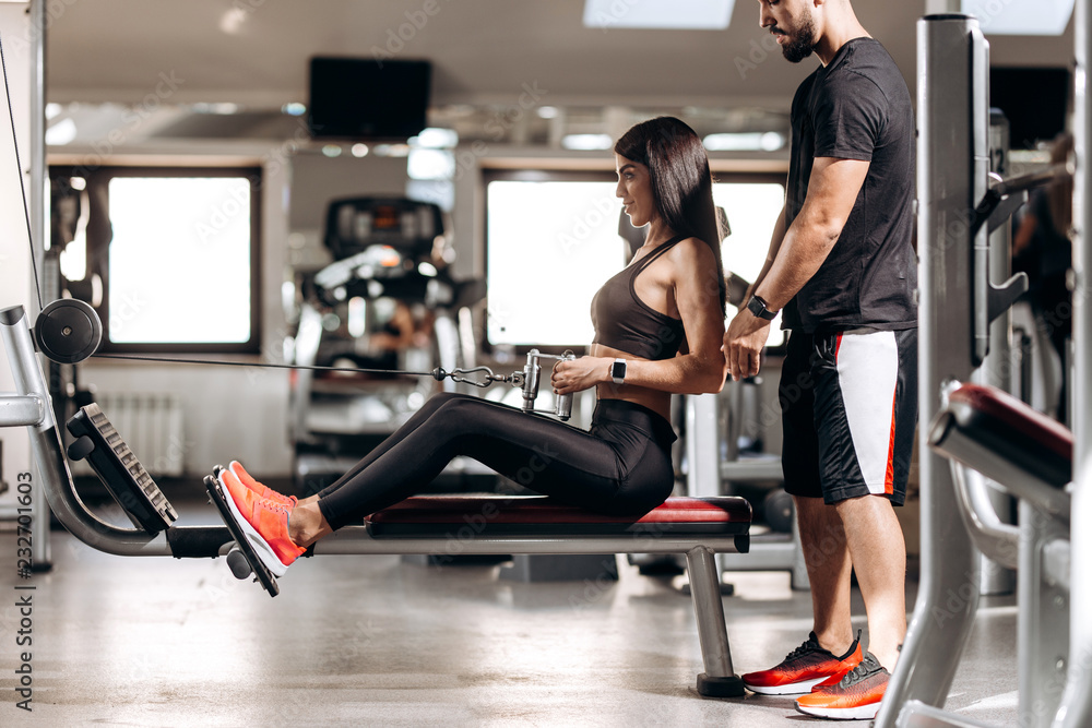 Beautiful slim girl dressed in black sport clothes is doing exercises for the abdominals  on a special exercise machine under the supervision of a coach in the gym