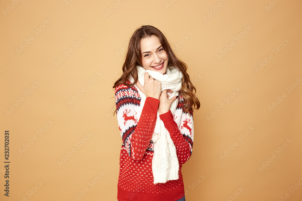 Beautiful girl dressed in a red and white sweater with deer and white knitted scarf stands on a beige background in the studio Beautiful girl dressed in a red and white sweater with deer and white knitted scarf stands on a beige background in the studio