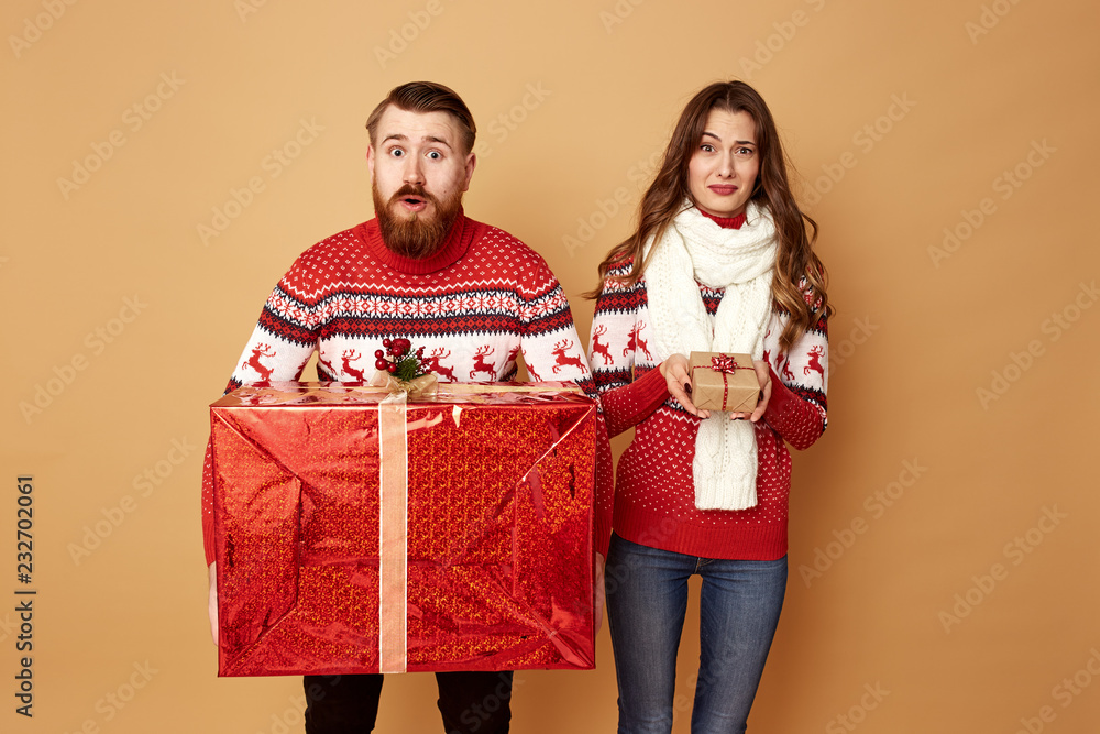 The girl holding a little gift surprisely and the guy holding a huge gift dressed in red and white sweaters with deer stand on a beige background in the studio The girl holding a little gift surprisely and the guy holding a huge gift dressed in red and white sweaters with deer stand on a beige background in the studio