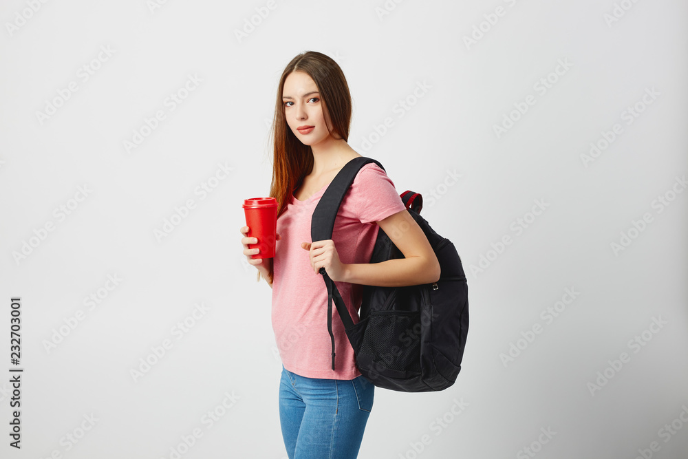 Nice girl with a black backpack on her shoulder dressed in a pink t-shirt and jeans holds a red plastic cup in her hand on a white background Nice girl with a black backpack on her shoulder dressed in a pink t-shirt and jeans holds a red plastic cup in her hand on a white background