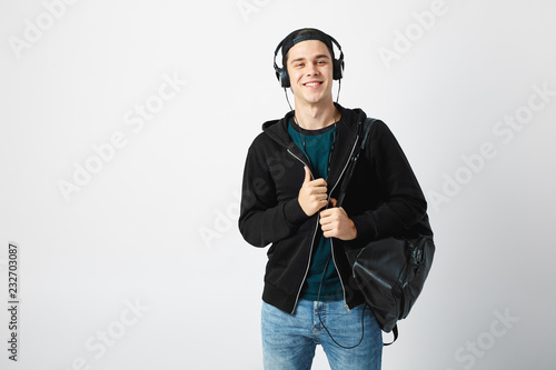 Happy guy with a black backpack on his shoulder and headphones on his head dressed in a dark t-shirt, jeans, sweatshirt and a cap uses mobile phone in the studio on a white background
