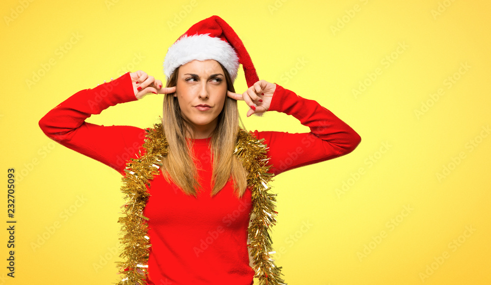 Blonde woman dressed up for christmas holidays covering ears with hands on yellow background