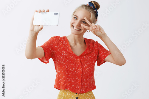 Unity of old and new. Portrait of stylish emotive and happy cute woman with fair hair in vintage oufit and headband showing peace sign at smartphone screen while smiling, holding phone to make selfie photo