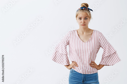 Intense confused and displeased young blonde wife in stylish striped blouse, holding hands on waist, raising one eyebrow with disbelief and doubt, being uncertaing about husband telling truth