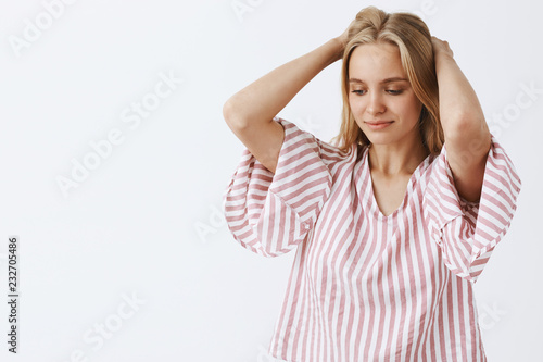 Waist-up shot of dreamy tender and feminine blonde caucasian woman in pink striped blouse holding hands behind head, gazing down with gentle slight smile thinking or imaging something