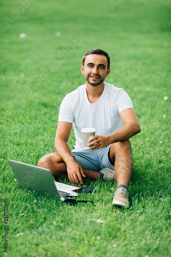 Young man sitting on the ground and working on laptop drinking coffee in park