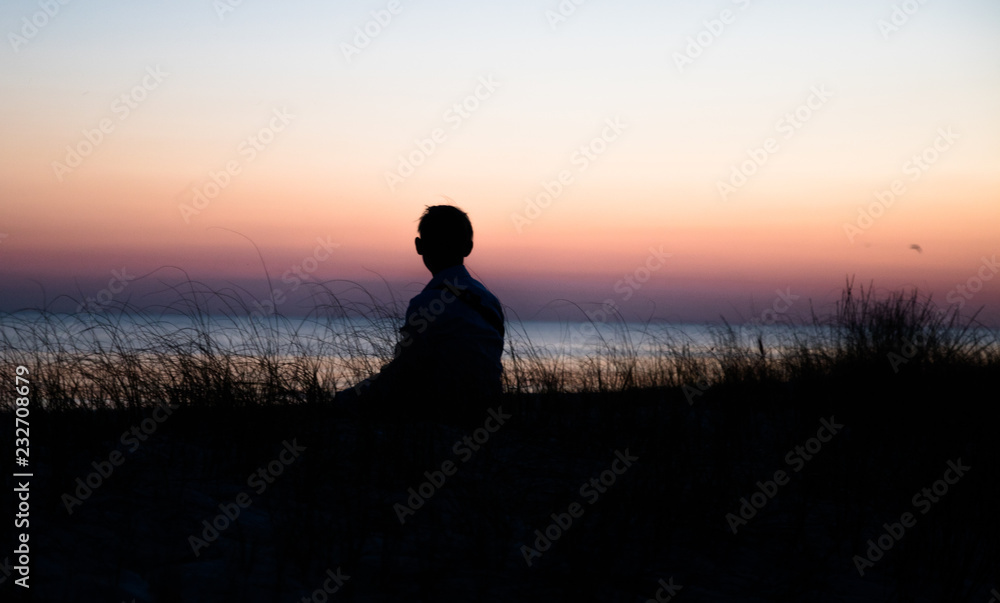 a boy sitting in the grass on the beach and admiring sunset