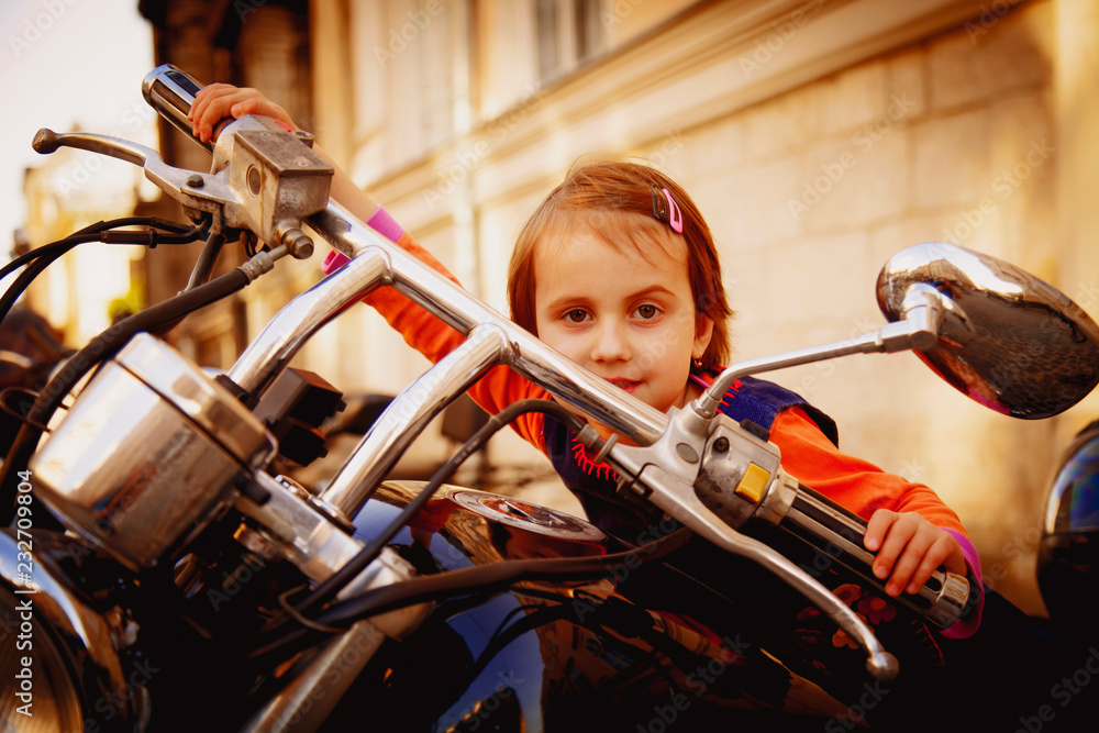 Humorous portrait of cute little biker child girl sitting on a vintage custom motorcycle as a symbol of freedom, adventure and travel.