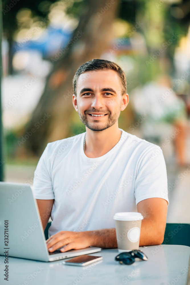 Outdoor portrait of young smiling man sitting at cafe table with laptop open enjoying summer time outside Outdoor portrait of young smiling man sitting at cafe table with laptop open enjoying summer time outside