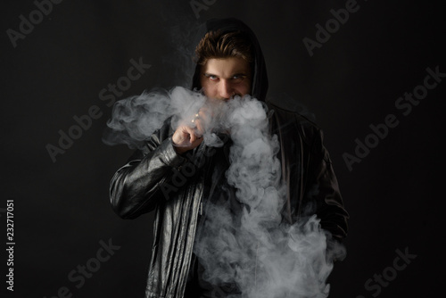 Man with vaping mod exhaling steam at black studio background. Bearded guy smoking e-cigarette to quit tobacco. Vapor and alternative nicotine free smoking concept