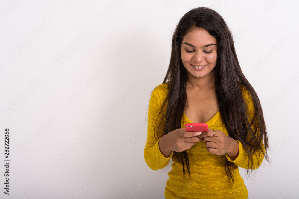Studio shot of young happy Indian woman smiling while using mobi Studio shot of young happy Indian woman smiling while using mobi