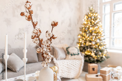 vase with cotton branches. Light living room with Christmas tree. Comfortable sofa, high large Windows