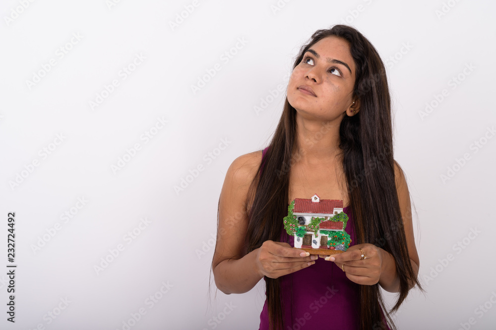 Studio shot of young Indian woman thinking while holding house f Studio shot of young Indian woman thinking while holding house f