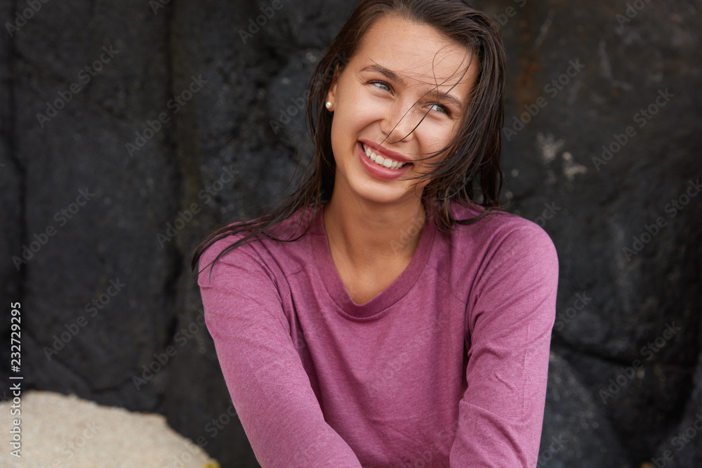Smiling glad Caucasian woman looks happily aside, being in high spirit after unforgettable rest on beach, poses at cliff, dressed in casual purple sweater, has positive expression. Summer time concept Smiling glad Caucasian woman looks happily aside, being in high spirit after unforgettable rest on beach, poses at cliff, dressed in casual purple sweater, has positive expression. Summer time concept