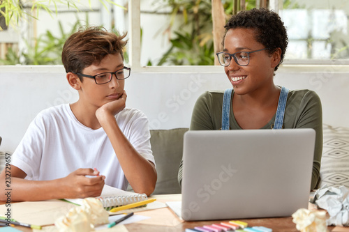 Self education and e learning concept. Satisfied black female volunteer tries to explain her strategy to young learner, watch attentively video in laptop computer. Tutor teaches skilled student photo