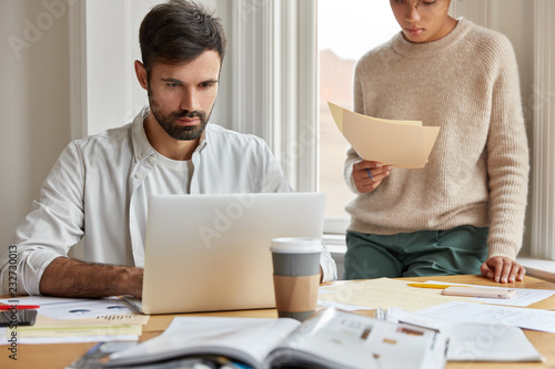 Indoor shot of serious man wants to deposite money in bank, reads job description on laptop computer, has much paperwork, unrecognizable dark skinned woman stands in background, develop market
